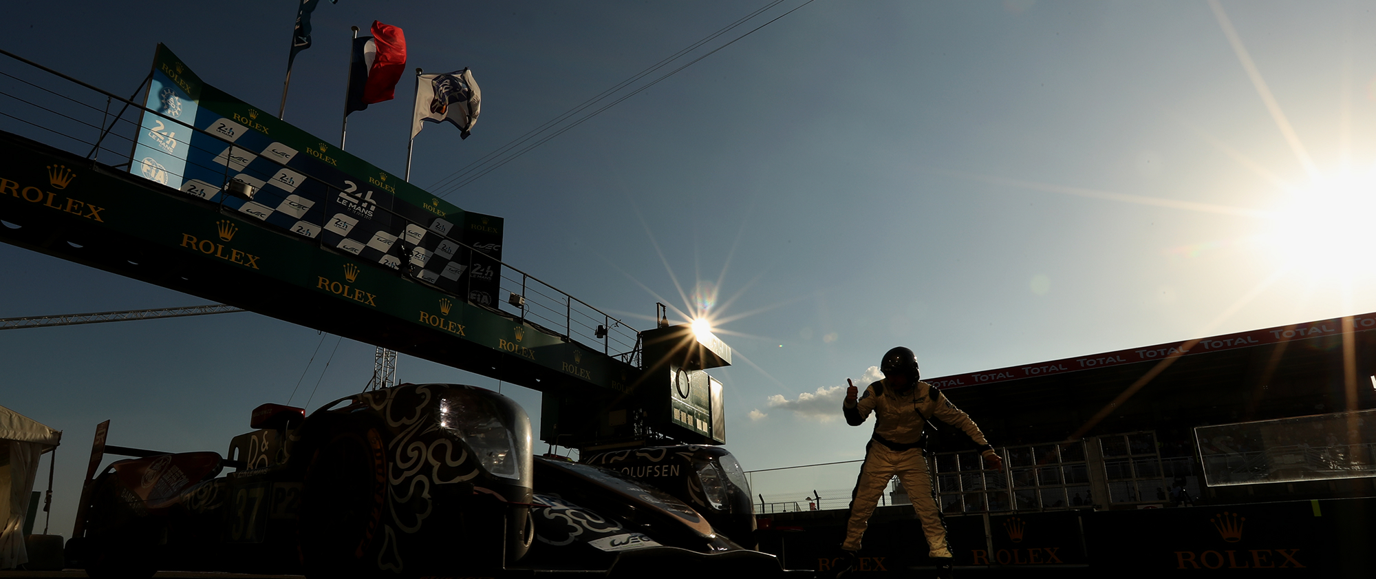 Le Mans 24 Hours finish line with race cars and crew silhouetted under the Rolex-branded podium at sunset